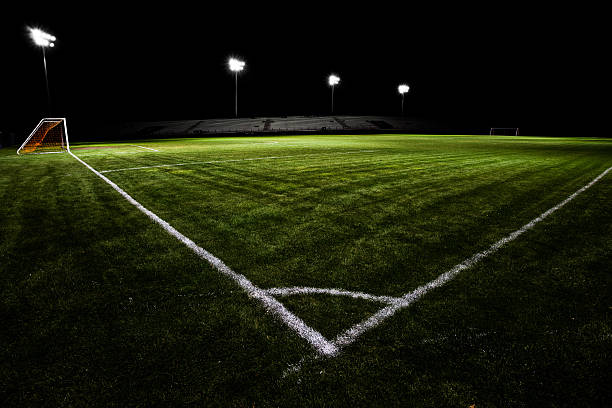 Groomed soccer field or soccer pitch at night.  Real, green grass, stadium style lighting and dark skies isolate the field from its surroundings.  The field is empty with no players.